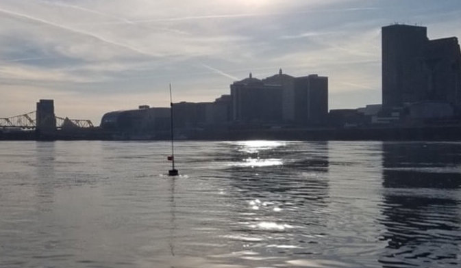 Waterway with bridge and buildings in background and floating buoy with pole and buoy node affixed to it in foreground