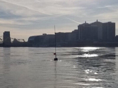 Waterway with bridge and buildings in background and floating buoy with pole and buoy node affixed to it in foreground