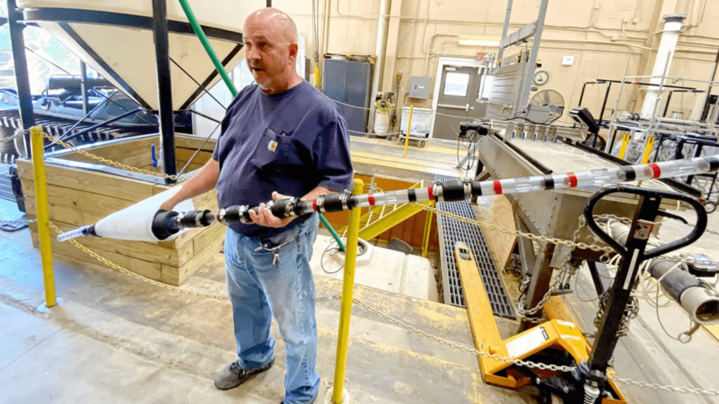 A man stands in a warehouse holding a long segmented clear rod with a conical mesh filter at its end.