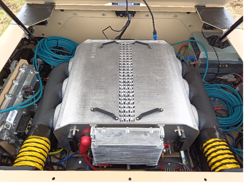 A gray box sits on worktable. On the left and right sides are three circular openings with black pipes extending outward and connected to each other. Yellow hoses extend from each pipe. On the closest end is a louvered block with red wires extending. Other ports are visible.