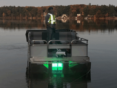 Front view of a pontoon boat with a UV reactor illuminated on the underside and the operator standing on the pontoon deck
