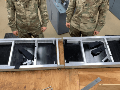 A portable modular scaling ladder, shown in two connected sections with straps and hardware exposed, is displayed on a workbench while three soldiers stand around it.