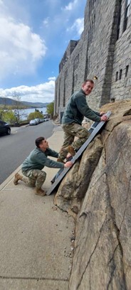 Two service members use a portable scaling ladder to climb a rocky embankment beside a stone building, with a road and parked cars visible below.