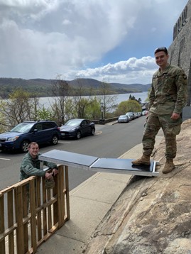 Two service members demonstrate a modular bridging ladder between a wooden railing and a rocky embankment, with a lake, parked cars, and mountains in the background.