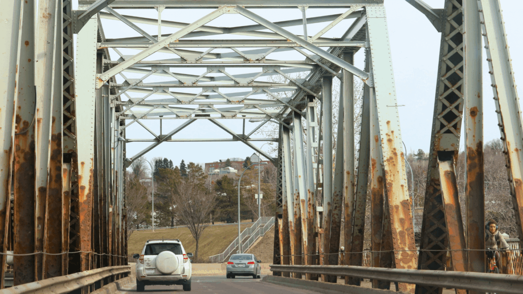 Two cars drive on a bridge with rust visible on its side structural beams