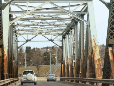Two cars drive on a bridge with rust visible on its side structural beams