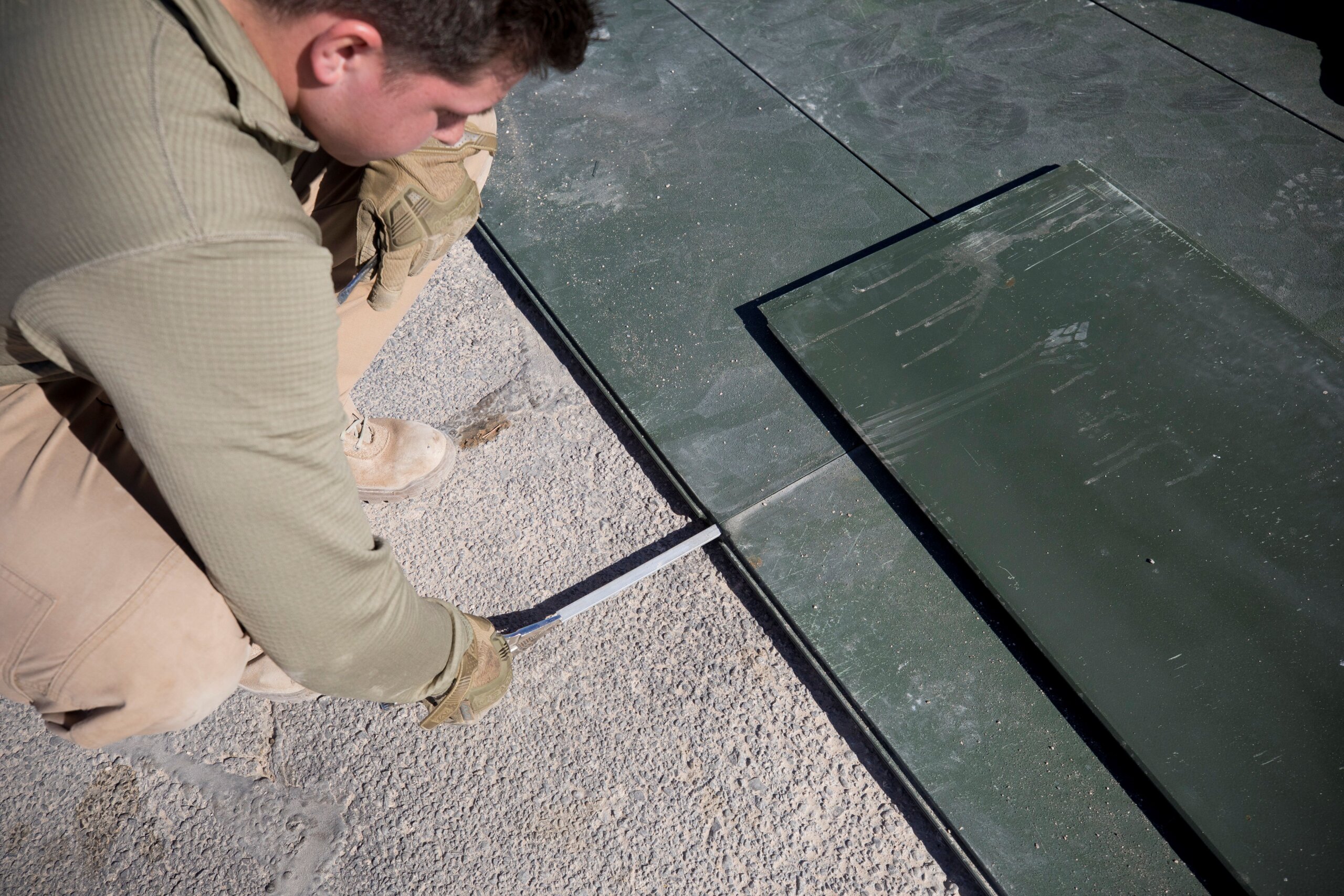 Worker with gloved hands uses pliers to pull a rectangular bar out of a hidden slot between two rectangular mats on an asphalt surface.